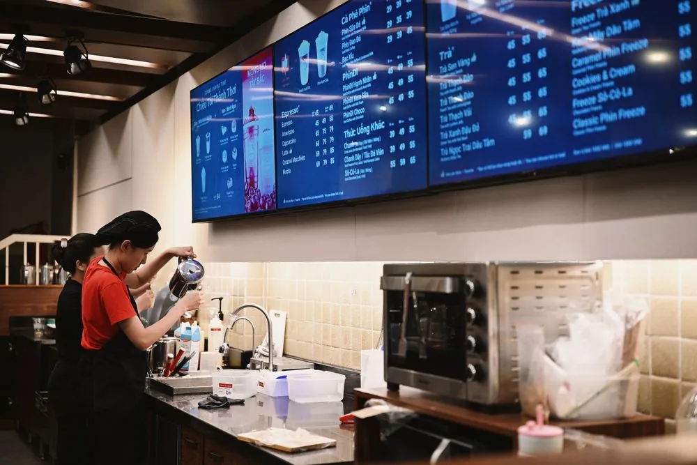 Three synchronised digital signage menu screens above a cafe counter with staff at work